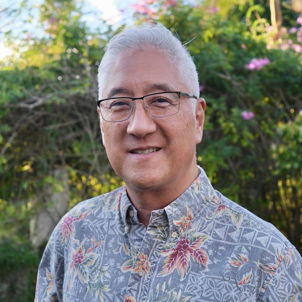 Dr. Sumi, a man with short white hair and glasses, smiles while standing in front of a garden.